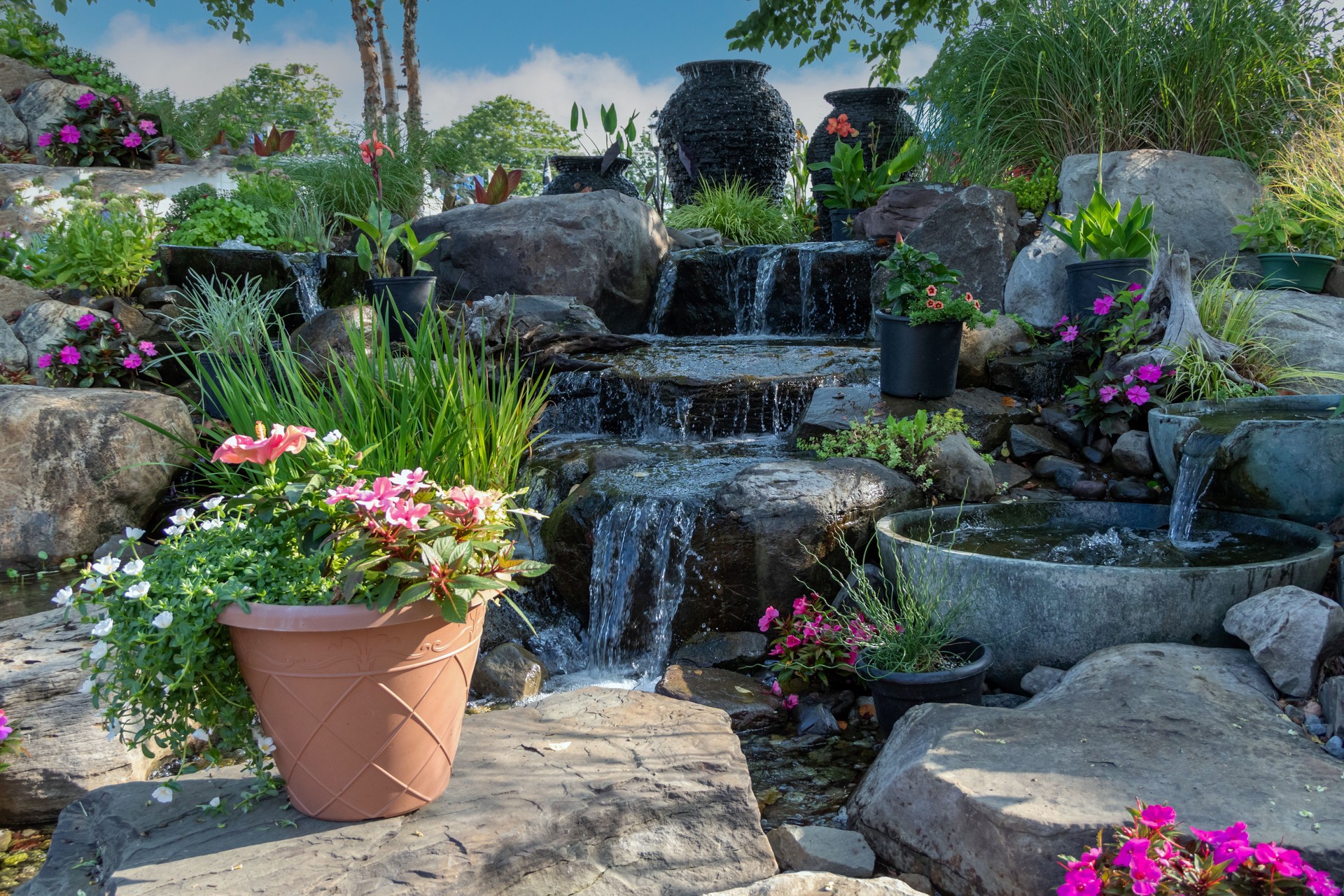 Waterscape fountain urns create a beautiful tranquil backyard oasis surrounded by bright pink and red flowers and ornamental grasses